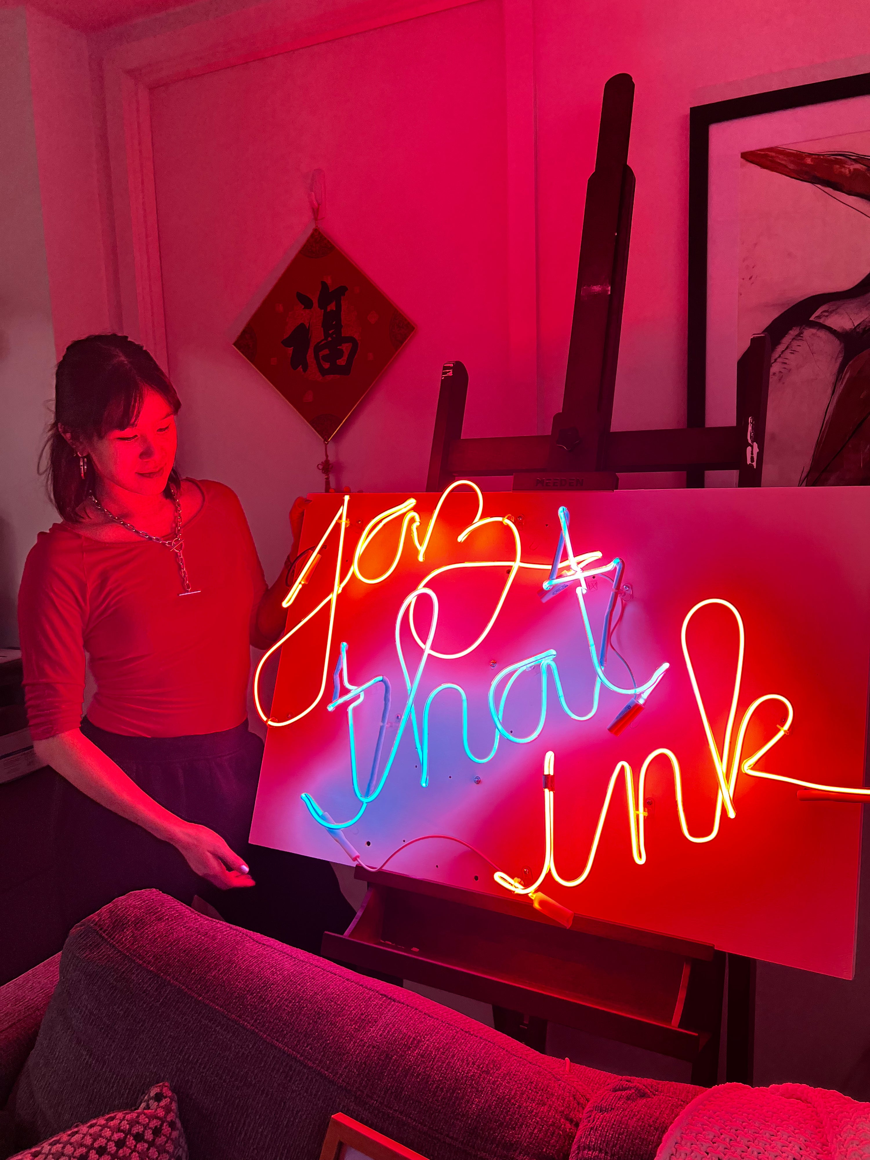 Bonnie standing in front of her lit neon sign which reads "Jaz that ink"